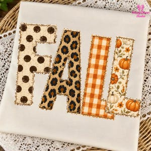 May include: A white t-shirt featuring the word FALL in large block letters. Each letter has a distinct pattern: the F is cream with brown polka dots, the A is leopard print, the L is orange gingham, and the second L features pumpkins.