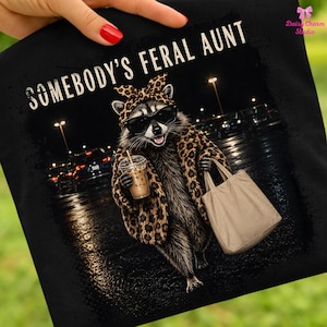 May include: A black t-shirt featuring a graphic of a raccoon wearing a leopard print headscarf, leopard print coat, and black sunglasses. The raccoon holds a plastic iced coffee cup and a beige tote bag. The text reads SOMEBODY'S FERAL AUNT.