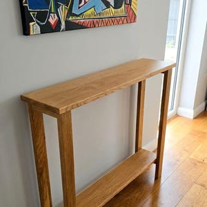 May include: A tall, narrow console table crafted from light brown wood with a visible grain pattern. The table features a rectangular top surface supported by four straight legs and includes a single lower shelf positioned between the legs.