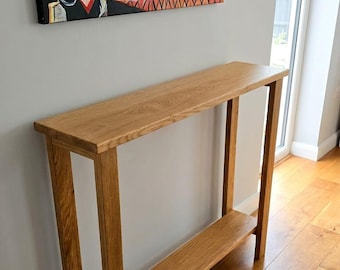 May include: A tall, narrow console table crafted from light brown wood with a visible grain pattern. The table features a rectangular top surface supported by four straight legs and includes a single lower shelf positioned between the legs.