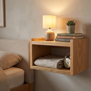 May include: A wooden floating bedside table with a lamp, books, and a small potted plant. A folded blanket and an alarm clock are inside the shelf. A framed botanical print hangs on the wall.