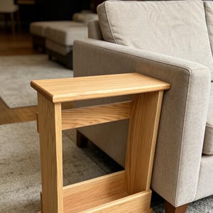 May include: A light-coloured wooden side table with a rectangular top and a lower shelf. The table is positioned next to a beige sofa, and the wood grain is visible. The table is approximately 61 cm tall.