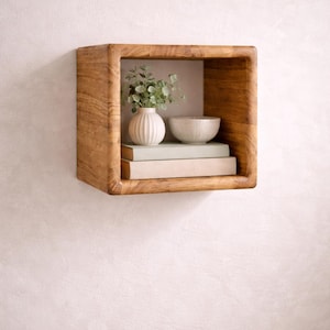 May include: A square wooden shelf with rounded edges is mounted on a white wall. Inside the shelf are two books, a small white vase with greenery, and a small, light-coloured bowl. The shelf is empty except for these decorative items.