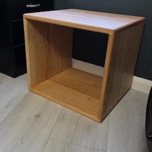 May include: A square, open-front wooden side table crafted from light-toned oak with a visible wood grain pattern. The piece features a smooth, continuous frame design with rounded edges, creating a hollow, box-like structure for storage.