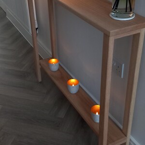 May include: A tall, narrow light wood console table with a lower shelf. On the top surface sits a white lamp with a conical shade and a glass reed diffuser. Three small, white, perforated votive candle holders with warm glowing lights sit on the lower shelf.