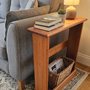 May include: A wooden side table in a reddish-brown hue. The table features a top surface with two books, a small potted plant, and a lamp. A woven basket is positioned below, holding newspapers.