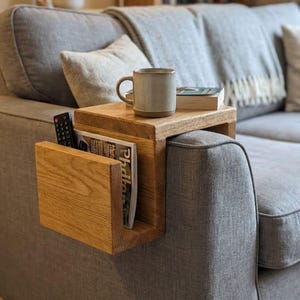 May include: Wooden sofa arm table with a magazine rack. A mug, book, and remote control rest on the table. The sofa is grey, and the table is made of light-coloured wood.