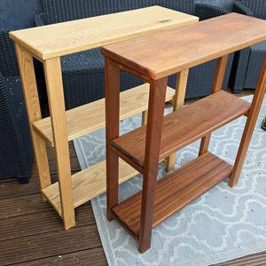 May include: Two rectangular wooden console tables with three tiers of shelving. One table is crafted from light-toned wood with a visible grain, while the other is constructed from a darker, reddish-brown wood. Both feature four vertical legs.