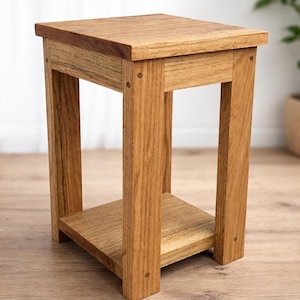 May include: A small, square wooden side table with a natural finish. The table has a top surface, a lower shelf, and four sturdy legs. The wood grain is visible, and the table is designed for indoor use. The table measures approximately 46 cm tall.