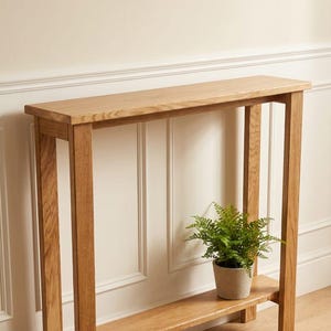 May include: A light oak console table with a lower shelf and four legs. A small potted fern sits on the lower shelf. A framed botanical print hangs on the wall above the table. The table is approximately 76 cm long.