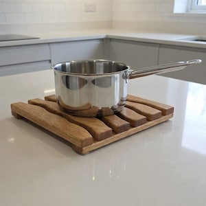 May include: A stainless steel saucepan with a long handle sits on a wooden trivet. The trivet is made of light brown wooden slats. The kitchen setting features a light-coloured countertop and cabinets. The trivet measures approximately 20 cm x 20 cm.