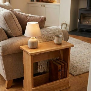 May include: A wooden side table with a lamp, books, and a mug. The table is made of light-coloured wood and has a shelf for storage. The lamp has a beige shade and a ceramic base. The mug is light brown.