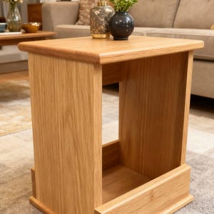 May include: A light oak square side table with a rectangular opening. The table has a smooth, natural wood grain finish. The table is 51 cm tall, 46 cm wide, and 46 cm deep. The table is in a living room setting.