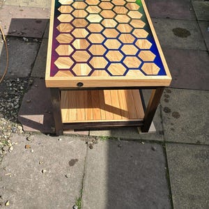 May include: A wooden coffee table with a distinctive honeycomb design. The tabletop showcases a pattern of wooden hexagons with colourful resin accents in purple, blue, and green. The table has a lower shelf and dark brown legs.