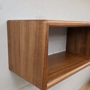 May include: A rectangular wooden shelf, finished in a warm brown, is mounted on a white wall. The shelf has a box-like design with rounded corners and a smooth, polished surface. The wood grain is visible.