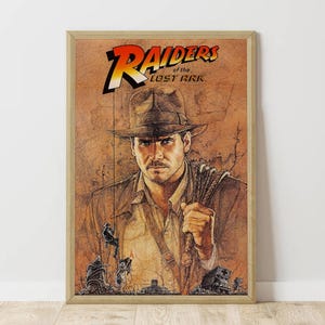 May include: A framed poster featuring the movie title "Raiders of the Lost Ark" with a portrait of the main character. The poster has a brown and orange color scheme, with the character wearing a fedora and holding a whip.