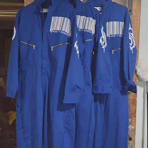 Puede incluir: Tres monos azules colgados de perchas blancas. Cada mono tiene una cremallera en la parte delantera, un gráfico de código de barras y diseños tribales blancos en las mangas. Los monos están hechos de una tela lisa de color sólido.