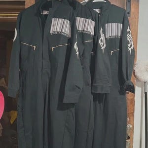 Puede incluir: Tres monos de trabajo verde oscuro colgando de perchas blancas. Cada mono tiene un código de barras y un diseño en las mangas. Los monos tienen una cremallera en la parte delantera y bolsillos. Los monos están hechos de un material duradero.