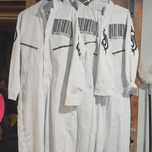 Puede incluir: Tres monos blancos colgados de una percha. Cada mono tiene un código de barras negro y cierre de cremallera. Un mono tiene un diseño tribal negro en la manga. Los monos están hechos de una tela duradera.