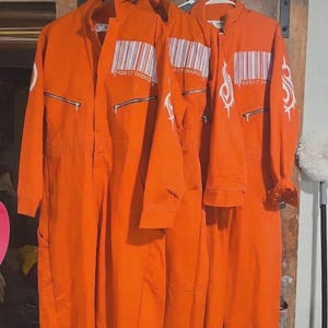 Puede incluir: Tres monos de trabajo de color naranja brillante colgados de perchas blancas. Cada mono tiene cierre de cremallera, bolsillos y un diseño de código de barras. Un mono tiene un gráfico blanco en la manga.