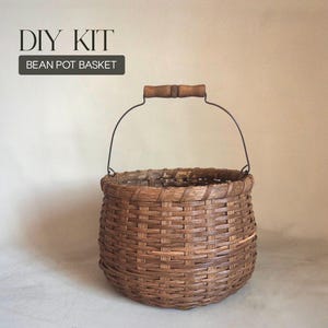 May include: A round woven brown basket with a dark metal wire handle featuring a wooden grip. The text DIY KIT BEAN POT BASKET is printed in the upper left corner. The basket has a wide, open top and a sturdy, interlaced construction.