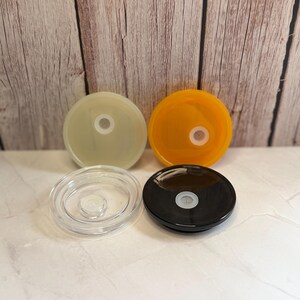 May include: Four colorful lids: translucent, orange, clear, and black. Each lid is round with a central hole. The lids are arranged on a white surface with a wooden background.