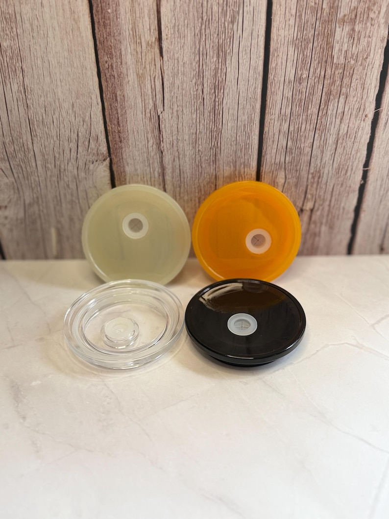 May include: Four colorful lids: translucent, orange, clear, and black. Each lid is round with a central hole. The lids are arranged on a white surface with a wooden background.