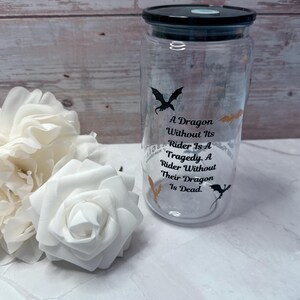 May include: A clear glass jar with a black lid, featuring dragon silhouettes and the quote "A Dragon Without Its Rider Is A Tragedy. A Rider Without Their Dragon Is Dead." White roses are in the foreground.