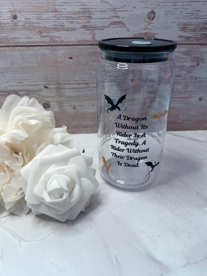 May include: A clear glass jar with a black lid, featuring dragon silhouettes and the quote "A Dragon Without Its Rider Is A Tragedy. A Rider Without Their Dragon Is Dead." White roses are in the foreground.