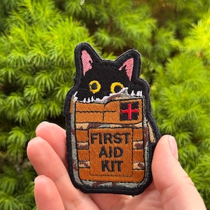 May include: Embroidered patch showing a black cat looking out of a brown first aid kit. The kit has a red cross and the words "FIRST AID KIT". The patch has a black border and is set against a green background.