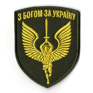 Op de afbeelding: Een schildvormige patch met een donkergroene achtergrond. Het ontwerp toont een engel met een zwaard en uitgespreide vleugels. Boven de engel staat de gele tekst "З БОГОМ ЗА УКРАЇНУ".