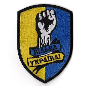 Op de afbeelding: Geborduurde patch in schildvorm, met een geel en blauw kleurenschema. Een vuist is in het midden, met de woorden "ВІЛЬНА УКРАЇНА!" op blauwe en gele banners. De patch heeft een zwarte rand.