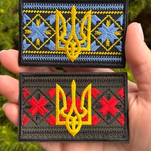 May include: Two rectangular embroidered patches featuring the Ukrainian Tryzub emblem in yellow. The top patch is black with blue geometric star patterns. The bottom patch is olive green with red geometric star patterns and black borders.