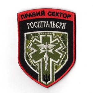 May include: Embroidered patch with a red border and black backing. The patch features text in white and a central design with a medical symbol, a dove, and a trident. The design is primarily green and white.
