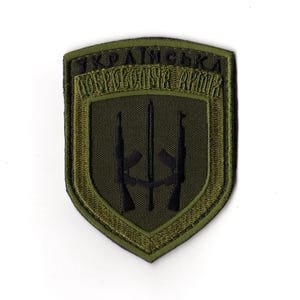 May include: An olive green embroidered patch with the text "УКРАЇНСЬКА ДОБРОВОЛЬЧА АРМІЯ" in black. The patch features a shield design with two black rifles and a trident symbol.