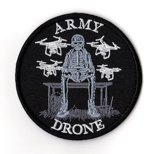 May include: Black circular patch with white embroidered text and imagery. The patch features the words "ARMY DRONE" and a skeleton figure seated on a bench, surrounded by four drone illustrations.