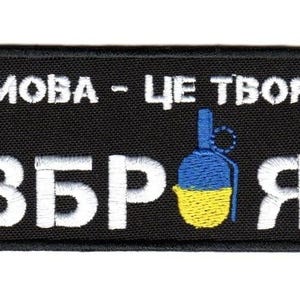 Puede incluir: Parche rectangular negro con texto bordado blanco y un gráfico de granada. El texto dice "MOBA - TSE TVOI ZBROYA" con una ilustración de granada azul y amarilla, probablemente con un tema político o militar.