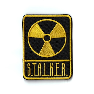 Pode incluir: Um patch bordado preto e amarelo com um símbolo de radiação amarelo sobre um fundo preto. A palavra "STALKER" é bordada em amarelo abaixo do símbolo. O patch tem uma borda preta e é retangular.