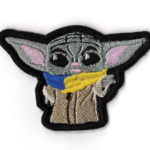 Puede incluir: Parche bordado con Grogu, un personaje con cabeza gris, orejas grandes con interior rosa y ojos negros grandes. El parche tiene un borde negro. Grogu lleva una túnica beige y una bufanda azul y amarilla.