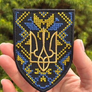Könnte beinhalten: Ein schildförmiger, bestickter Aufnäher mit schwarzer Stoffbasis und einem goldfarbenen ukrainischen Tryzub-Emblem in der Mitte. Der Aufnäher ist mit komplizierten gelben und blauen Kreuzstichmustern verziert, die das zentrale Dreizacksymbol umgeben.