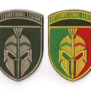 May include: Two embroidered patches with the text "INTERNATIONAL LEGION". One patch is green and tan, featuring a helmet and sword design. The other patch is red, green, and yellow, with the same helmet and sword design.