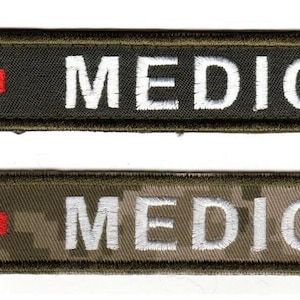 May include: Two embroidered patches with the word "MEDIC" in white block letters. Each patch has a red cross in the upper left corner. One patch is black with a green border, the other is camouflage with a green border.