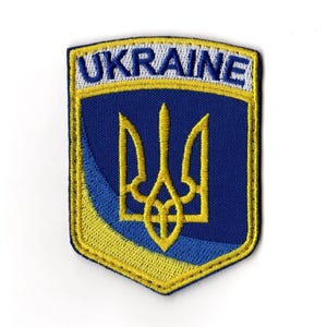 Puede incluir: Parche bordado con la palabra "UKRAINE" en blanco sobre un borde amarillo. El parche tiene un fondo azul con un emblema de tridente amarillo. El diseño tiene forma de escudo.