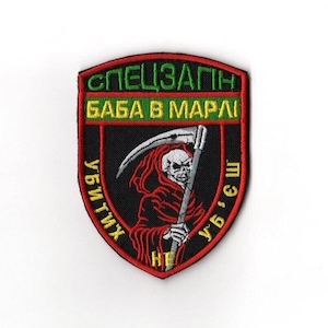 May include: Embroidered patch with a black background and a red, green, and white design. The patch features a Grim Reaper figure with a scythe and text in a foreign language. The patch is shield-shaped.