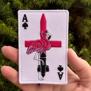 Puede incluir: Un parche bordado rectangular con fondo blanco y diseño de carta de juego. Muestra un flamenco rosa de pie sobre una bomba negra, colocado frente a una cruz roja. Las esquinas presentan la letra A y un símbolo de pica negro.