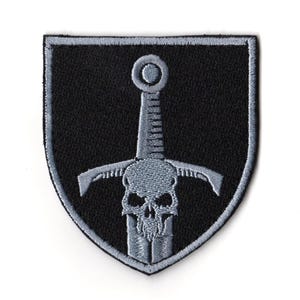 May include: A black and grey embroidered patch in a shield shape. The patch features a grey sword piercing a skull, with a grey border. The background is black.