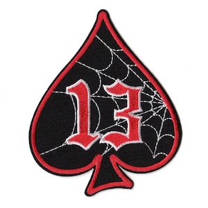 May include: A black patch in the shape of a spade with a red border. The number "13" is embroidered in red gothic font, with a white spiderweb design across the black background.