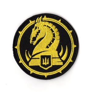 May include: Circular black patch featuring a yellow horse head design within a yellow border. The horse head faces left, with a shield shape beneath. The border has yellow triangular points.
