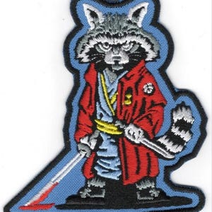 Puede incluir: Parche bordado con un mapache con abrigo rojo, camisa azul y fajín amarillo, sosteniendo una espada. El mapache tiene una expresión severa y está sobre un fondo azul claro con un borde azul oscuro. Diseño único.