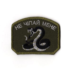 以下が含まれることがあります： 黒と白の刺繍が施されたヘビが特徴のオリーブグリーンのパッチ。パッチの上部には白い文字で「НЕ ЧІПАЙ МЕНЕ」と書かれており、デザインに大胆な印象を与えています。